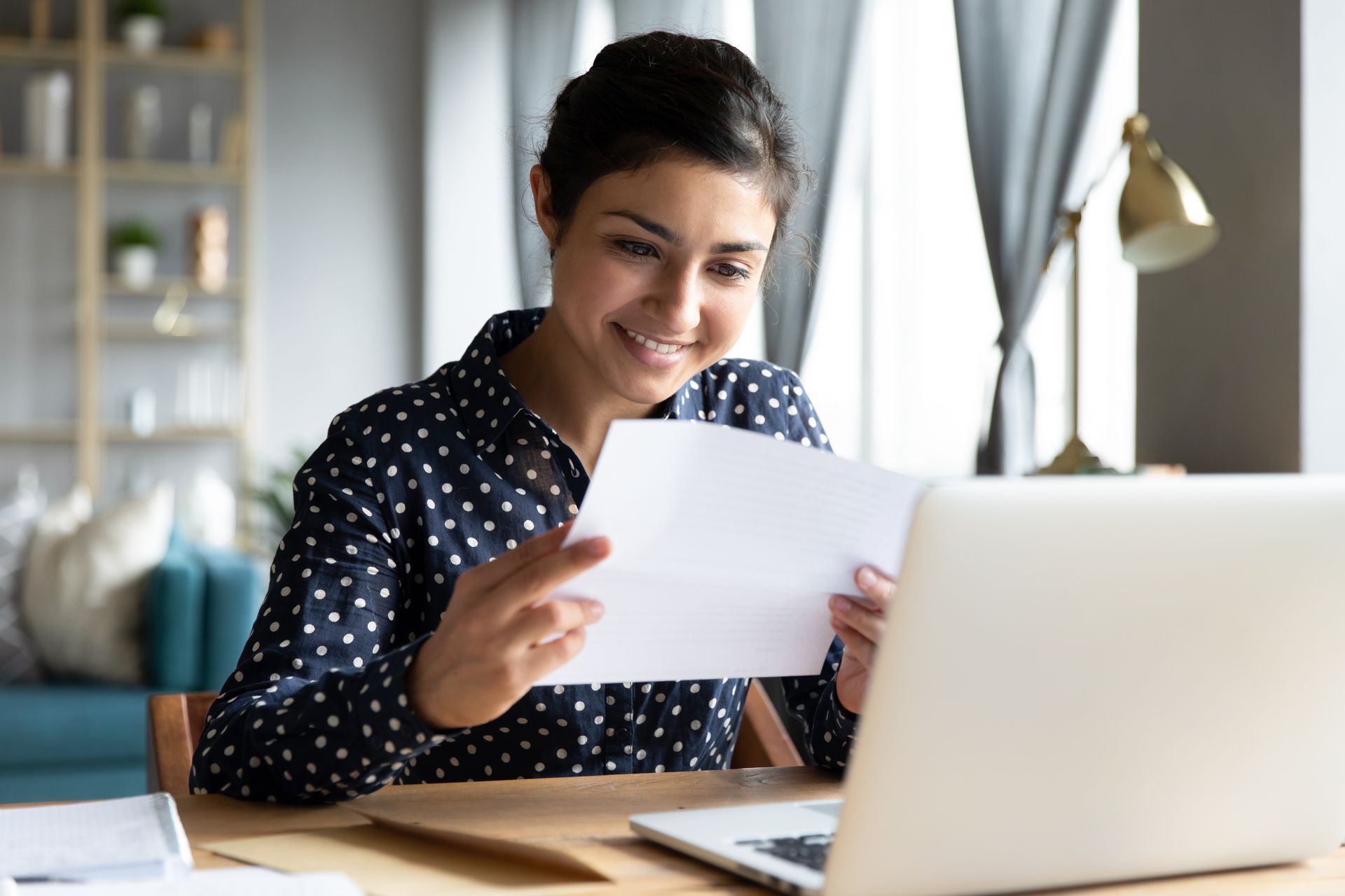 A woman is sitting at a desk holding a piece of paper in front of a laptop computer.