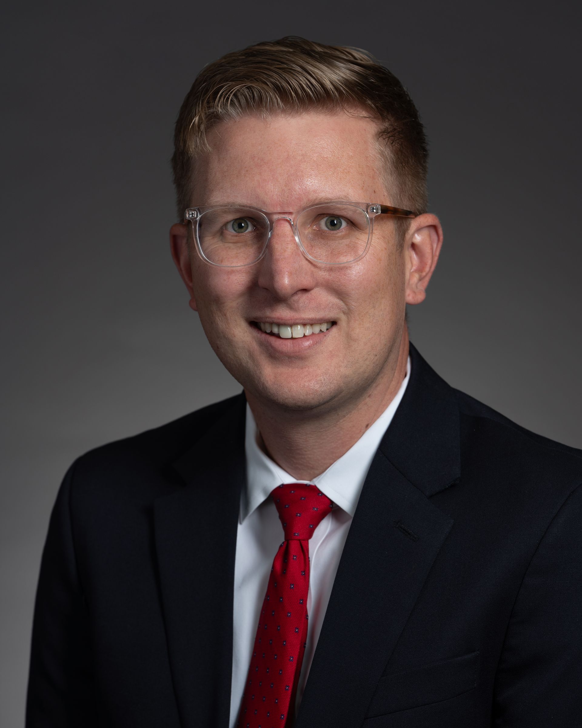 Man in suit and glasses, smiling. Red tie, against a gray backdrop.