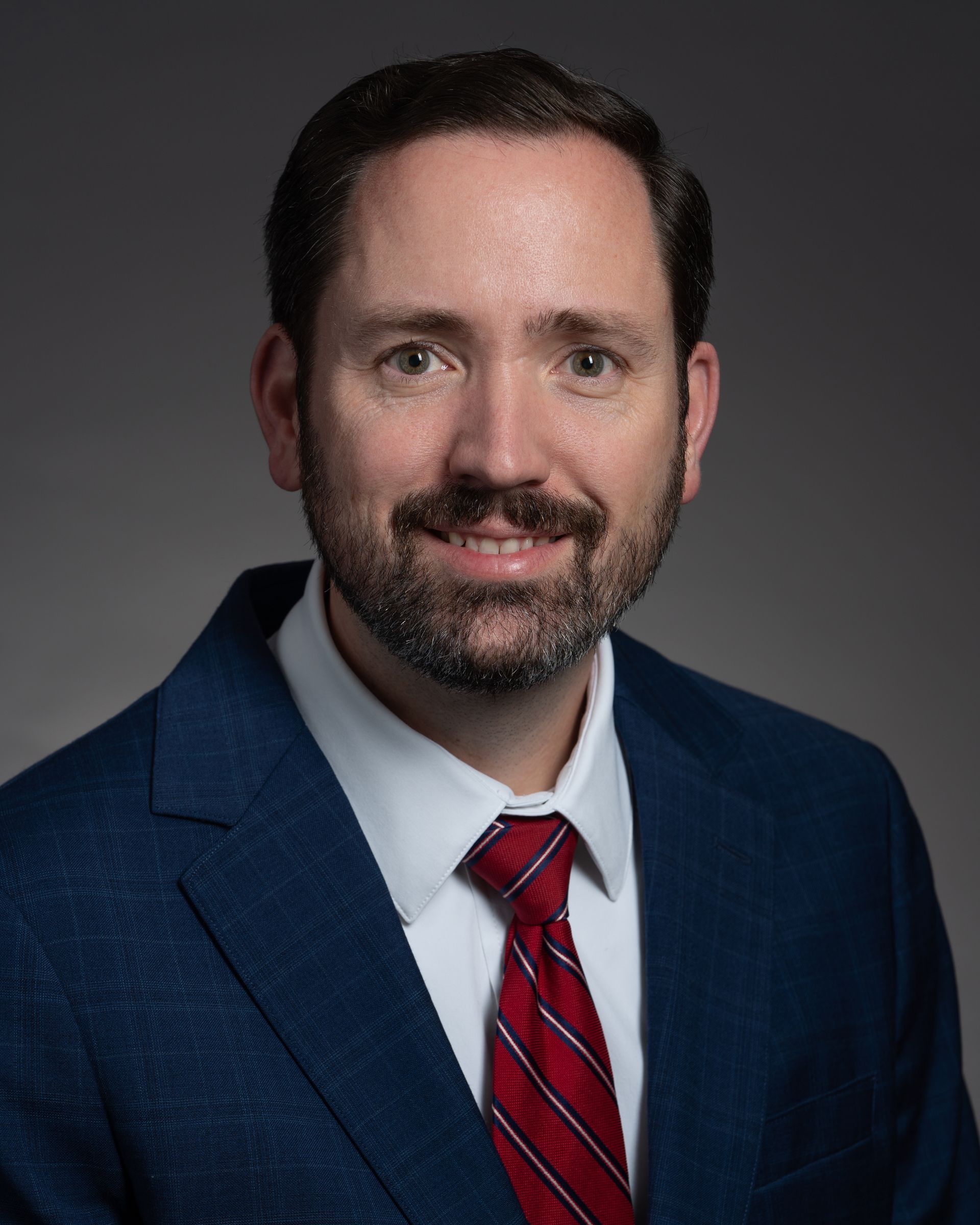 Man in a blue suit, white shirt, and red tie smiling. Gray background.
