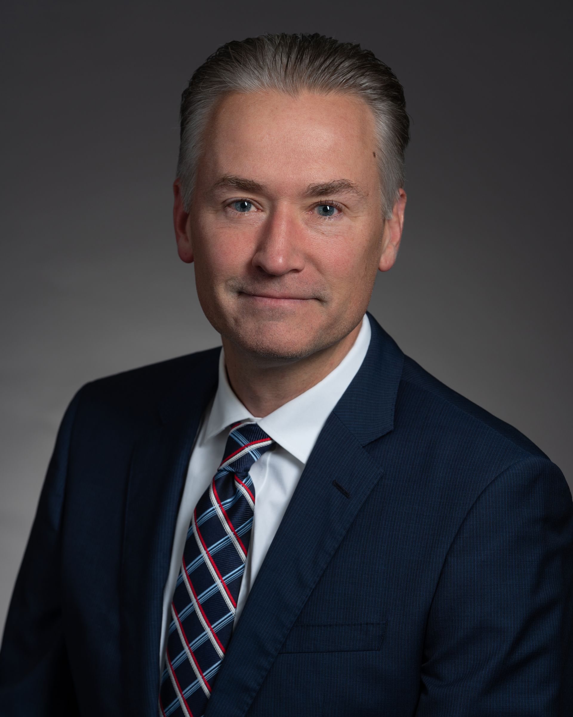 Man in suit, smiling, with a patterned tie, set against a neutral gray background.
