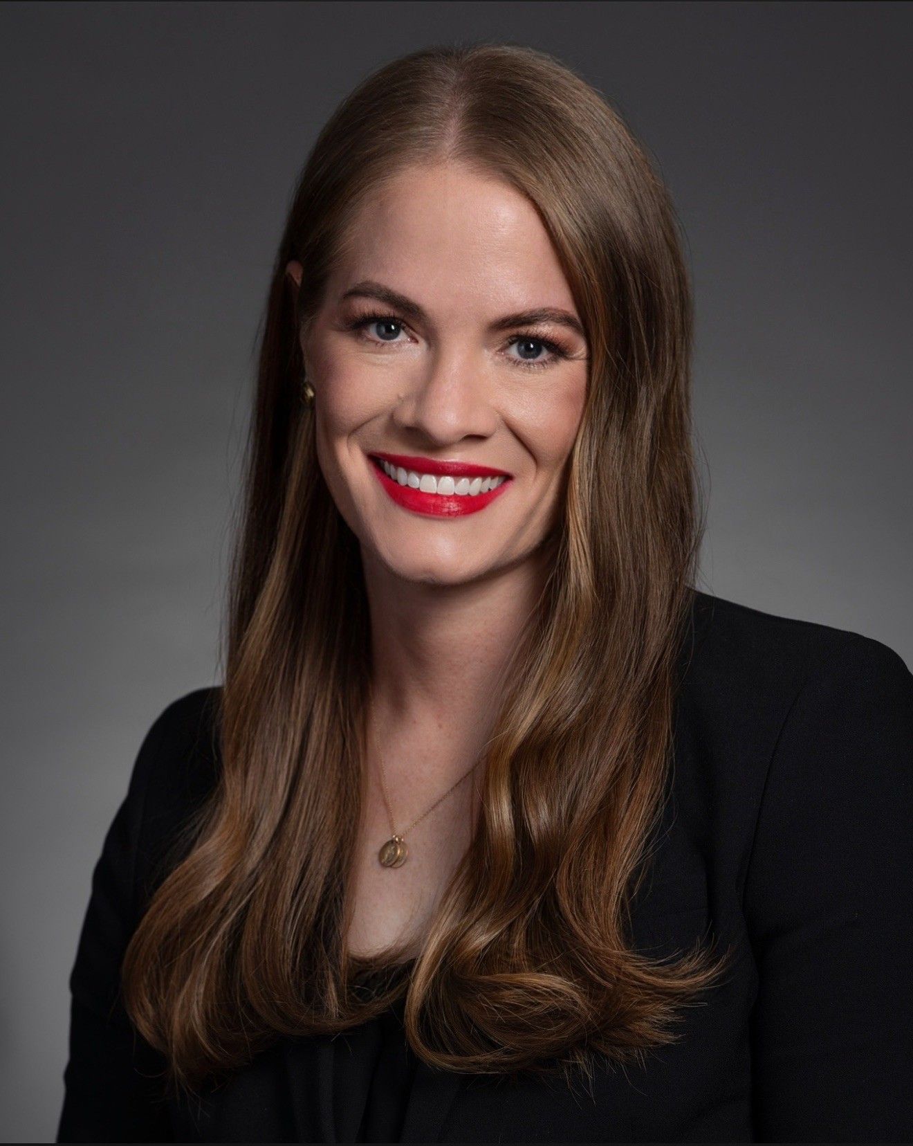 Woman with long brown hair, wearing black jacket and red lipstick smiles at the camera.