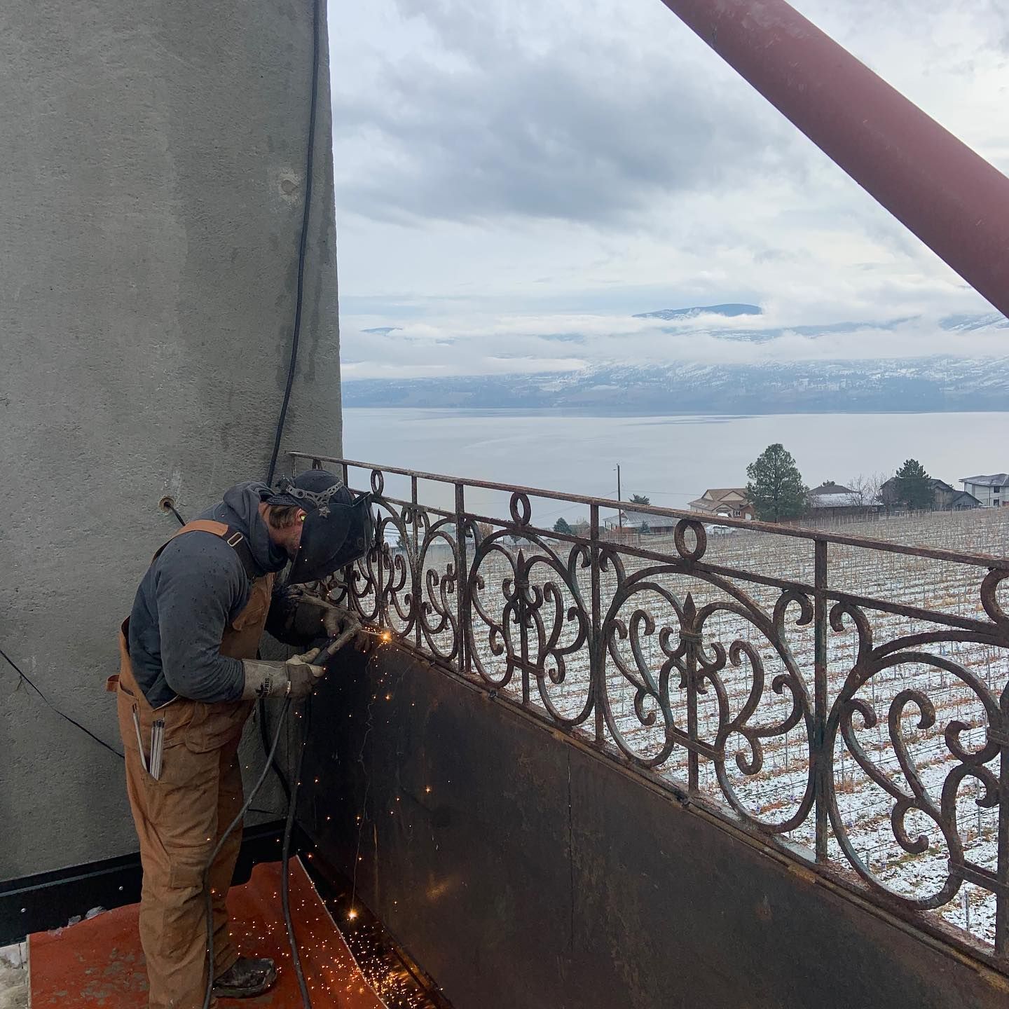 A man is welding a metal railing on a balcony overlooking a lake.