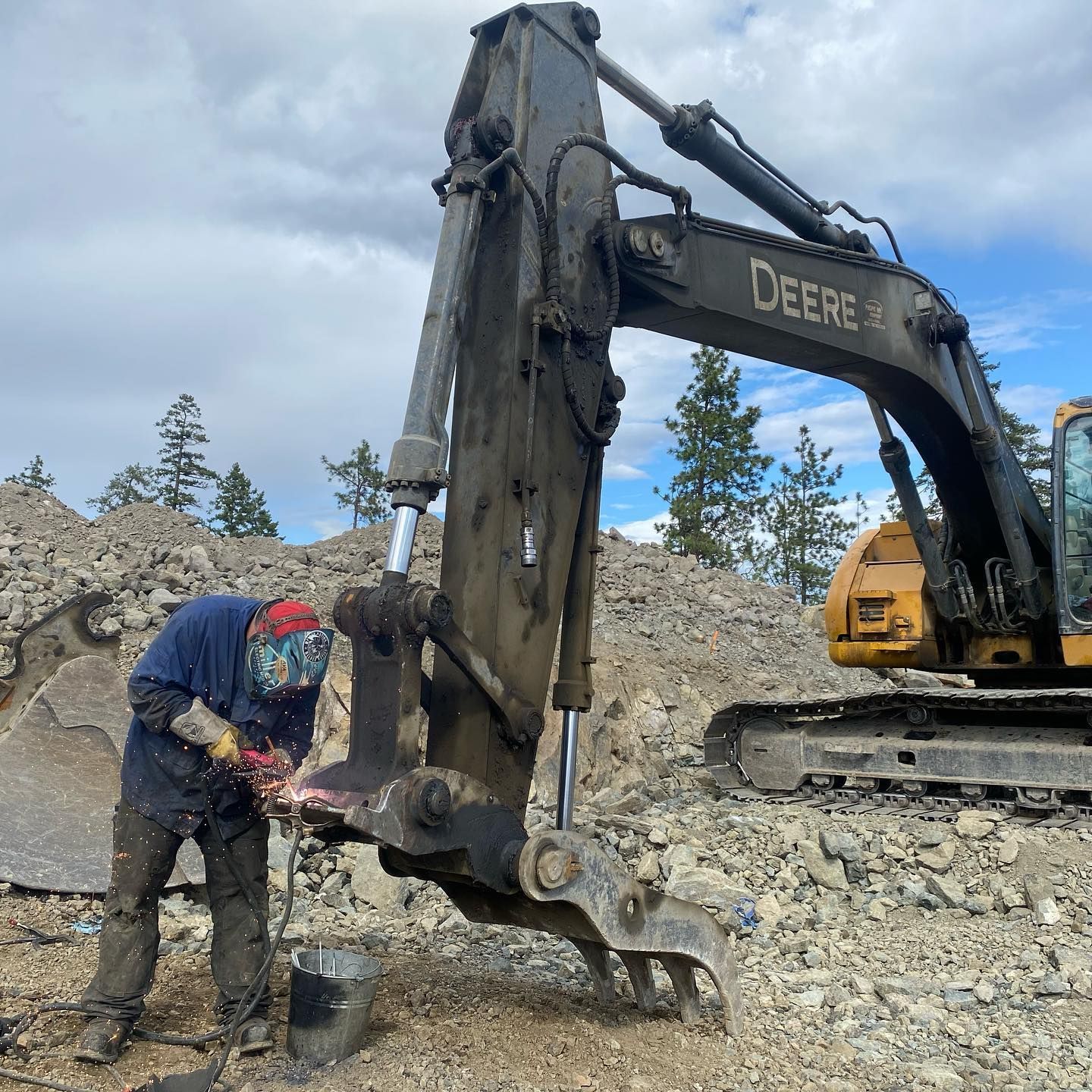 A man is welding a piece of equipment in front of a deere excavator.