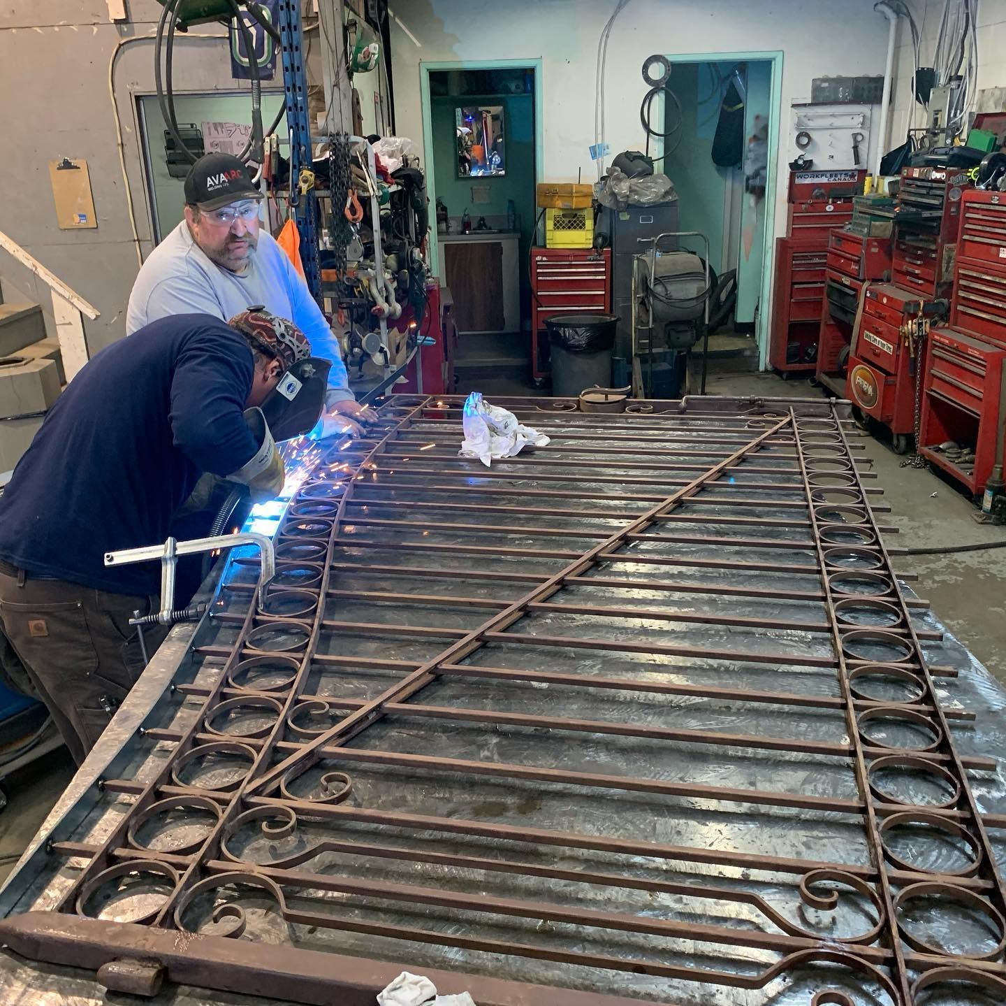 Two men are welding a metal fence in a workshop.