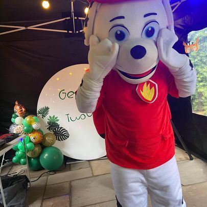 a mascot wearing a red shirt and white pants is standing in front of balloons .