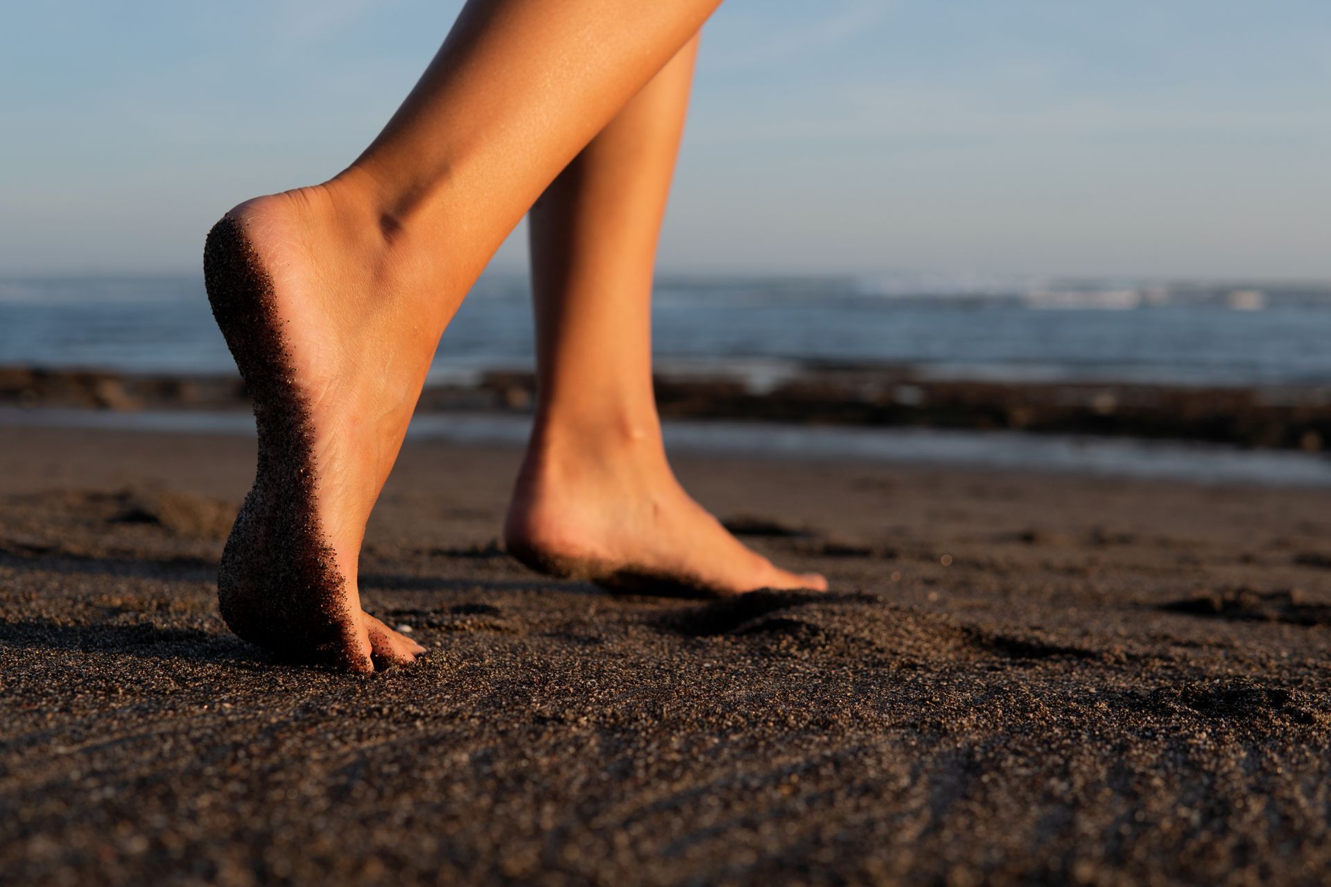 Bare feet walking on dark sand at a beach near the ocean.