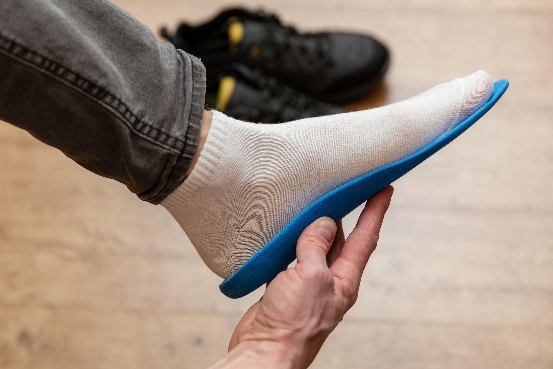 A hand holds a blue shoe insole against the sole of a sock-covered foot, with black sneakers in the background.