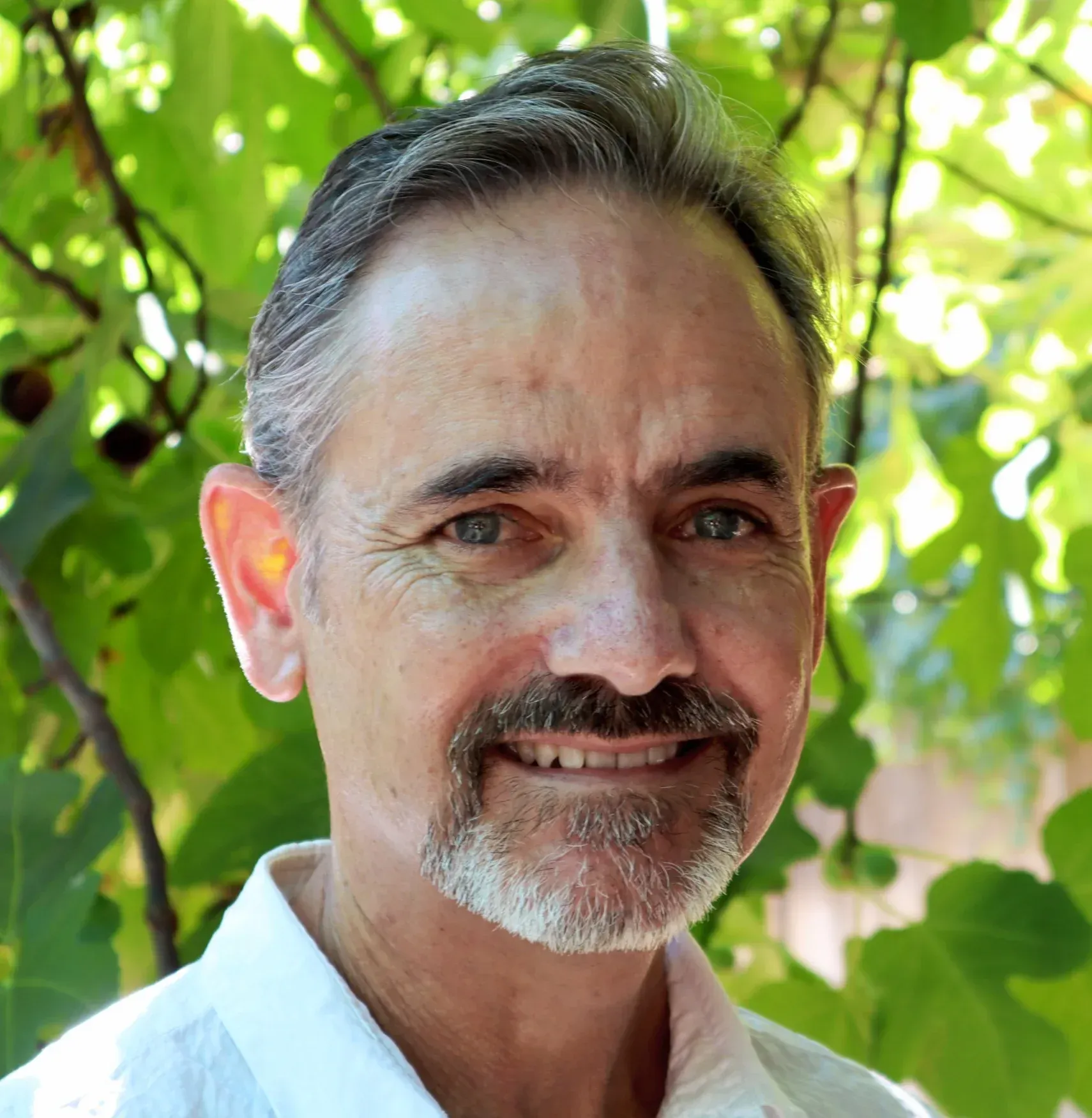 Man with graying hair and beard smiles in front of leafy green background.