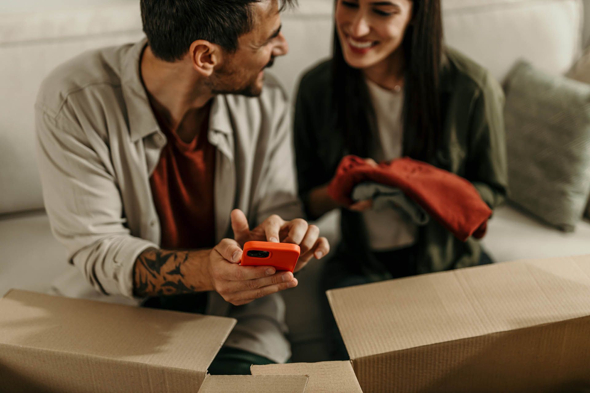 A man smiles at the woman sitting next to him on a couch as he checks his smartphone