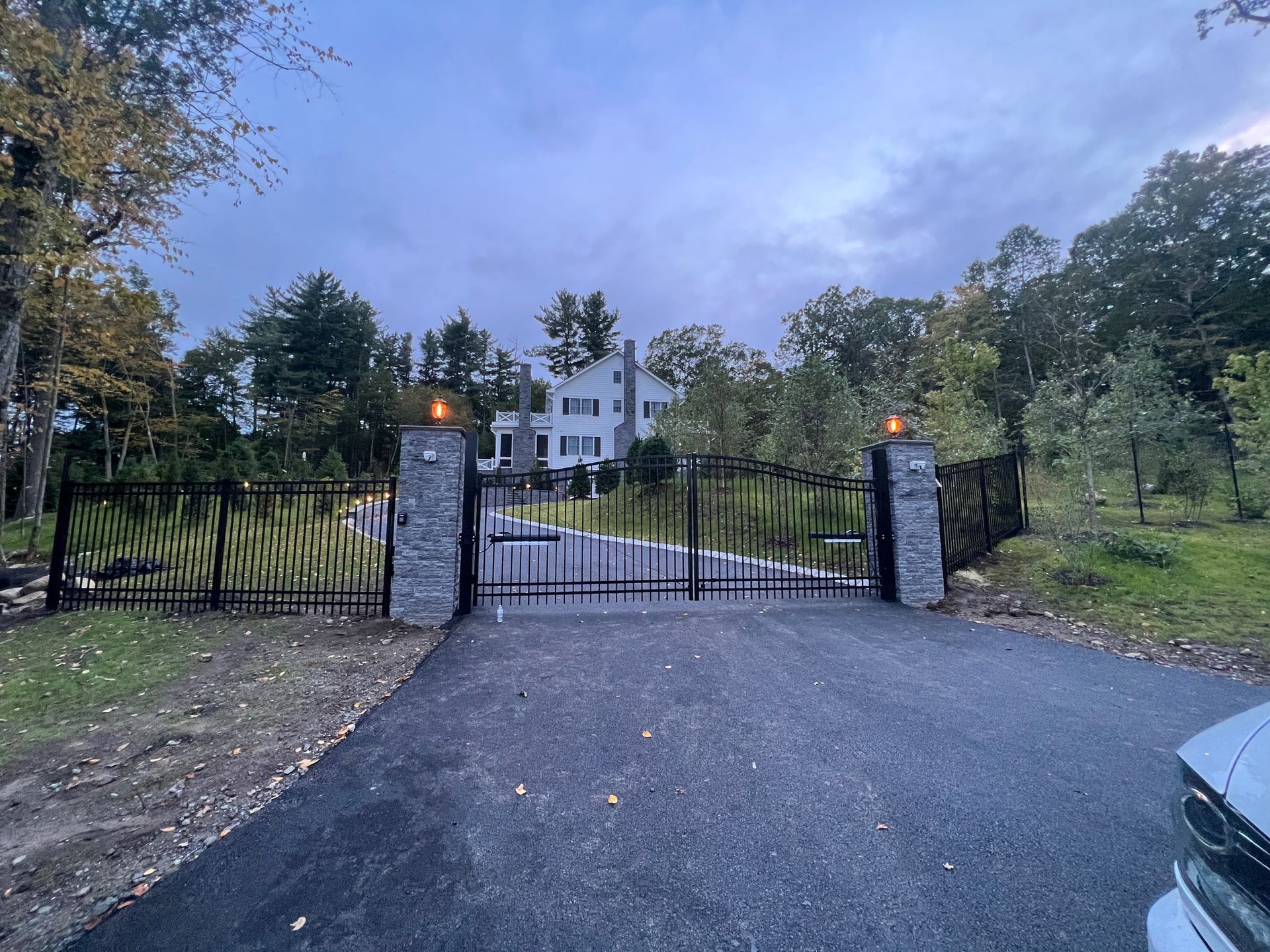 A black metal gate closes off a paved driveway leading to a large white house on a sunny day.