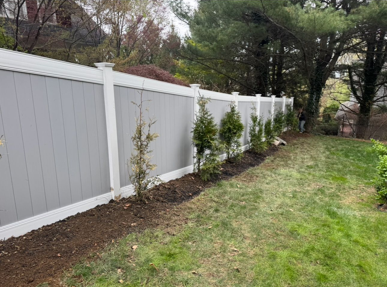 A long brown vinyl privacy fence stands in a grassy backyard with bare trees against a sunset sky.