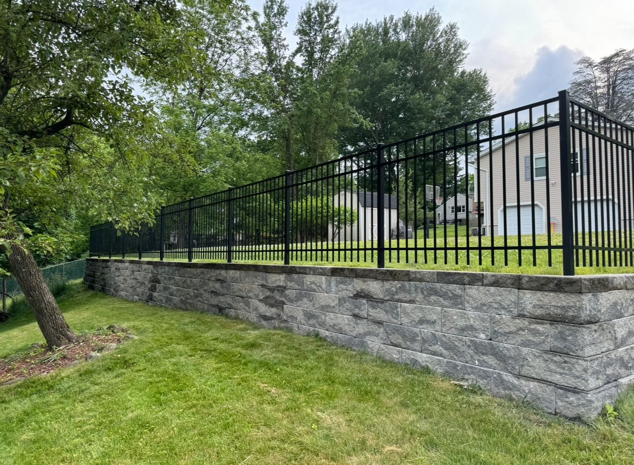 A tall, black horizontal-slat privacy fence stands along a wooded property line on a sunny day.