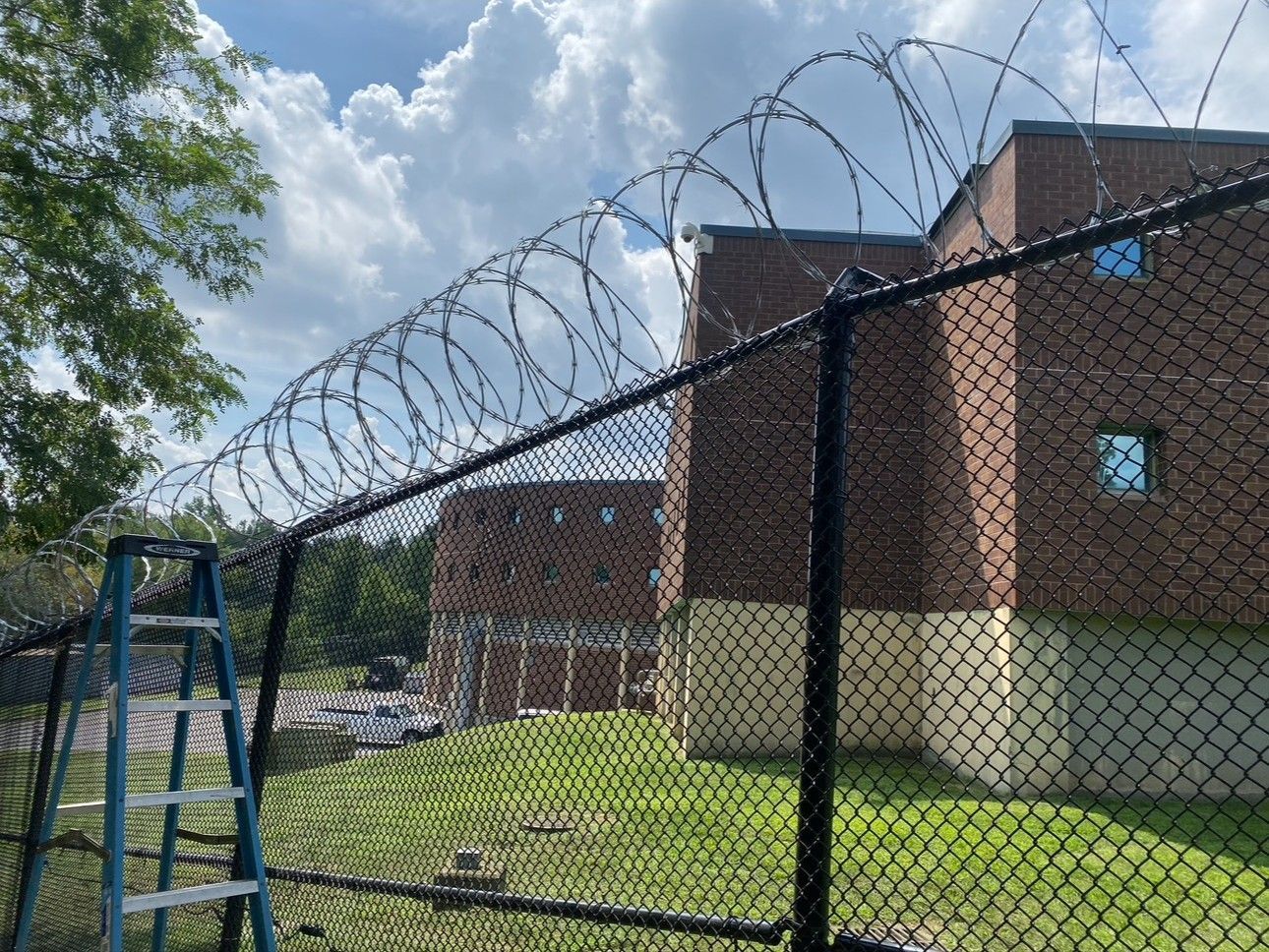 A brick building surrounded by a chain-link fence topped with barbed wire, with a blue stepladder in the foreground.
