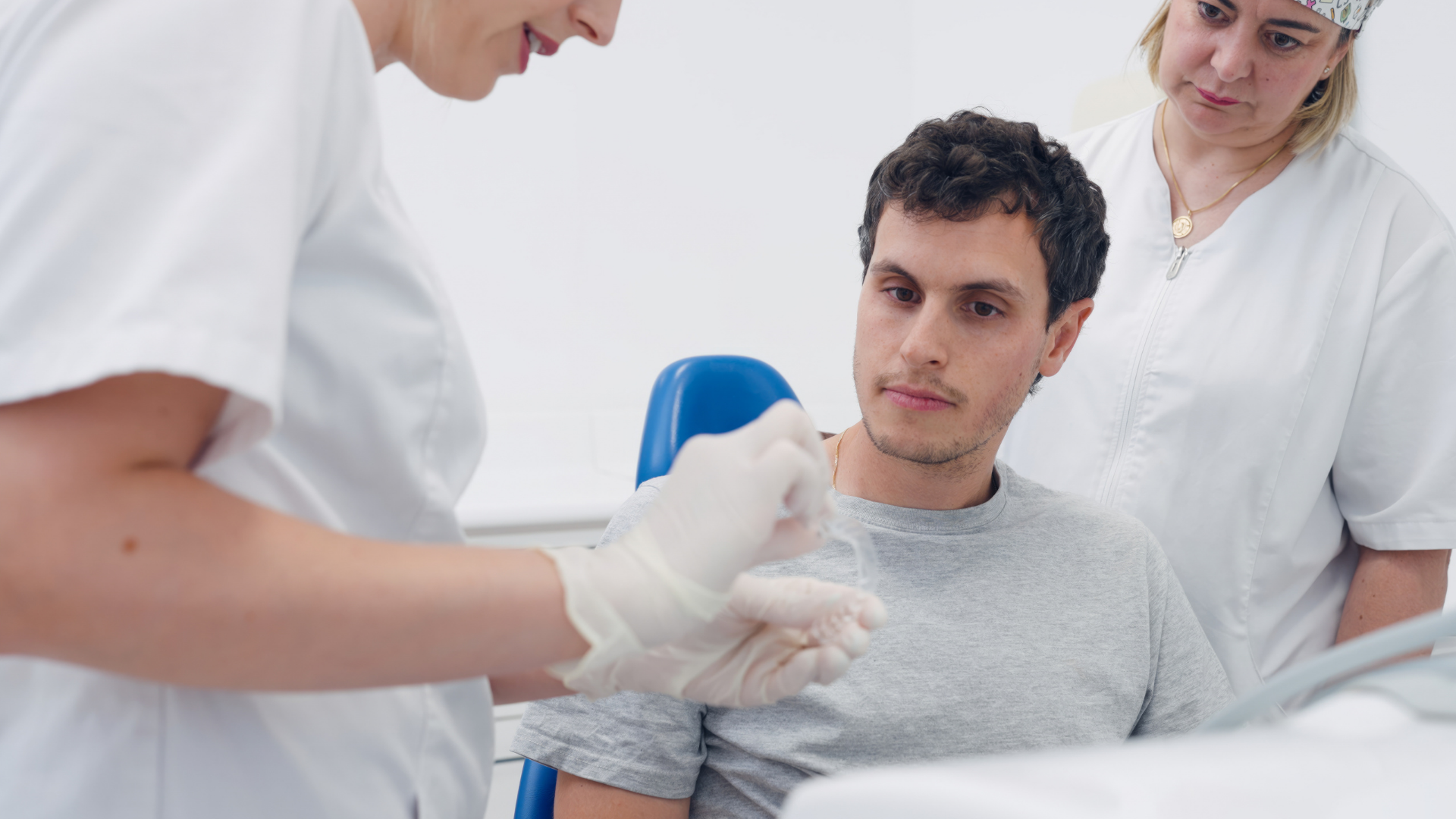 Two medical professionals in white uniforms consult with a patient sitting in a chair during a clinical appointment.