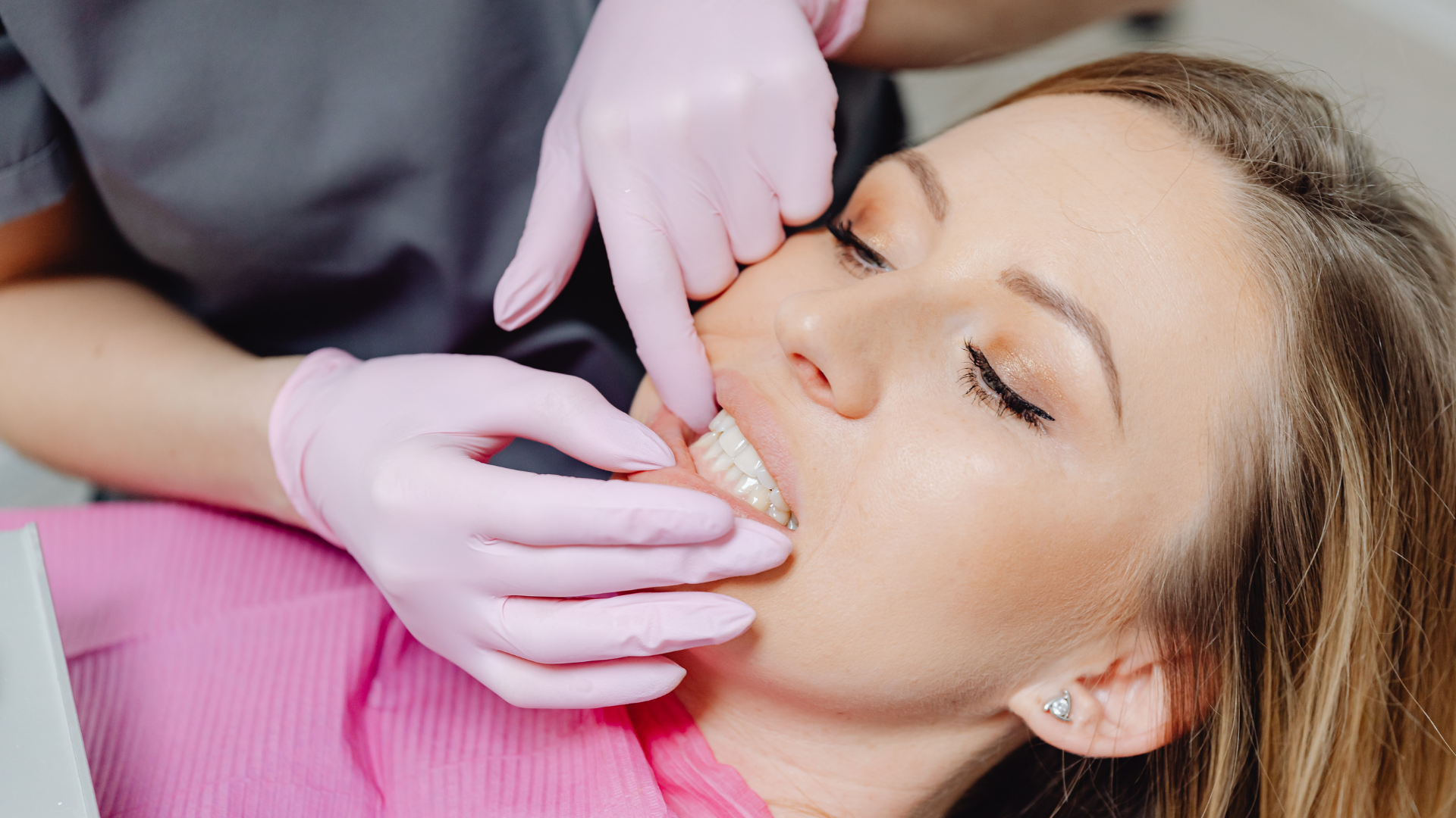 Dentist fitting clear dental aligner in patient's mouth. Pink gloves, neutral background.