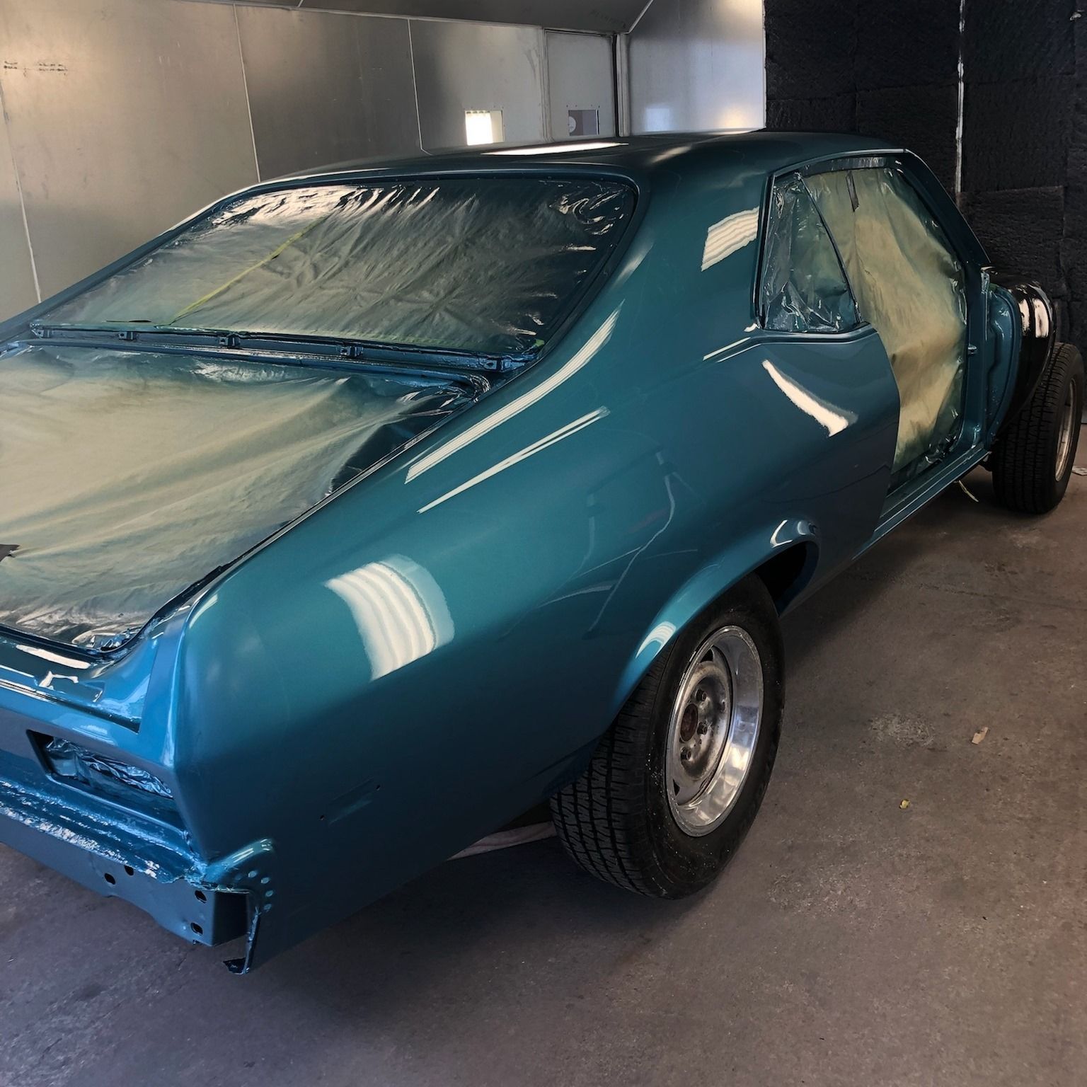 Blue classic car in a paint booth, partially masked, being painted.