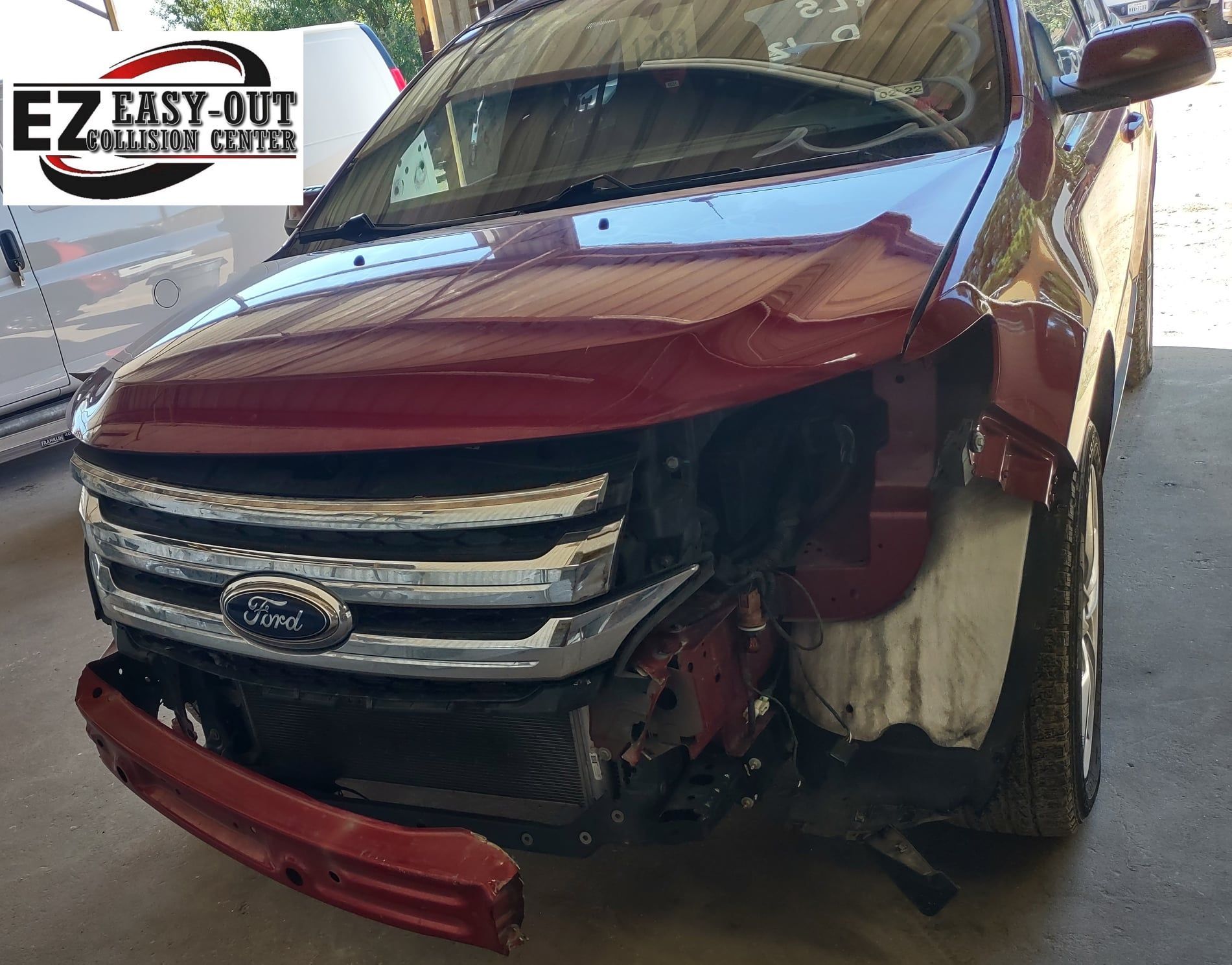 Damaged red Ford Edge missing front bumper and fender at a collision repair center.