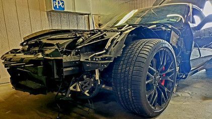 Black car with severe front-end damage, likely after a collision. The door is open, and a garage setting.