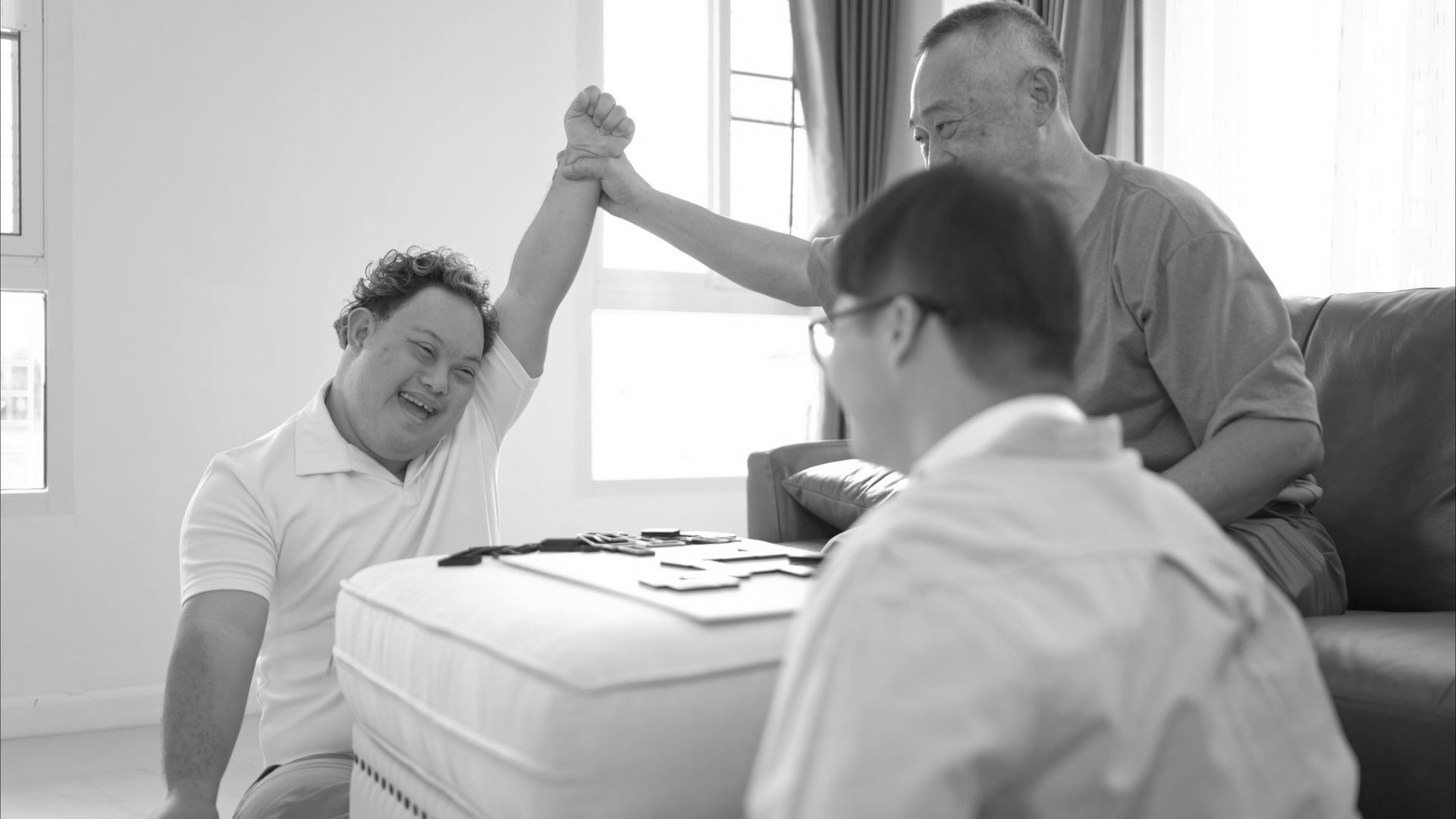 Three people playing a game. One raises arm in victory, cheered by another. Dominoes on a low table.