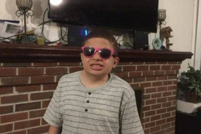 Boy wearing pink sunglasses, gray striped shirt, standing in front of a brick fireplace, looking at camera.