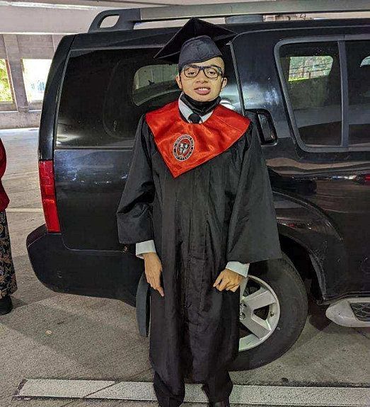 Person in graduation gown stands in front of a black SUV. They wear glasses, a black cap, and a red and black stole.