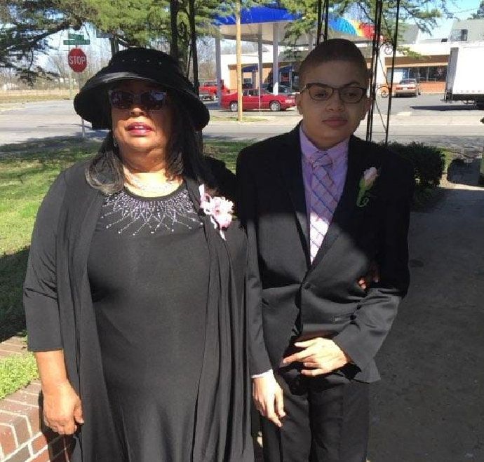 Woman and person in formal wear standing outdoors. Buildings, gas station in the background.