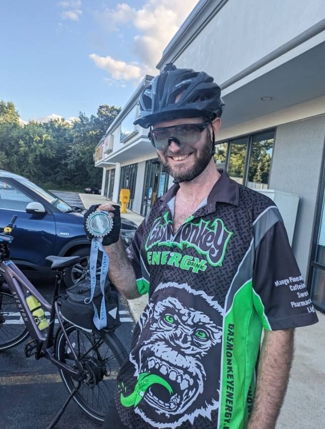 Cyclist in black-and-green jersey holding a bike light outside a shop, smiling at the camera.