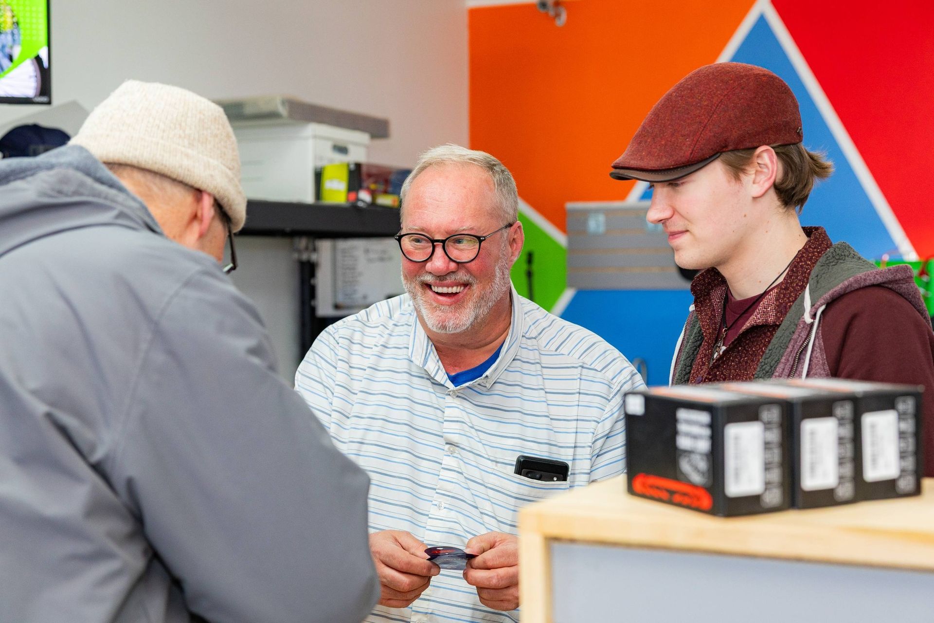 Three people talking at a counter in a colorful indoor shop or workshop.