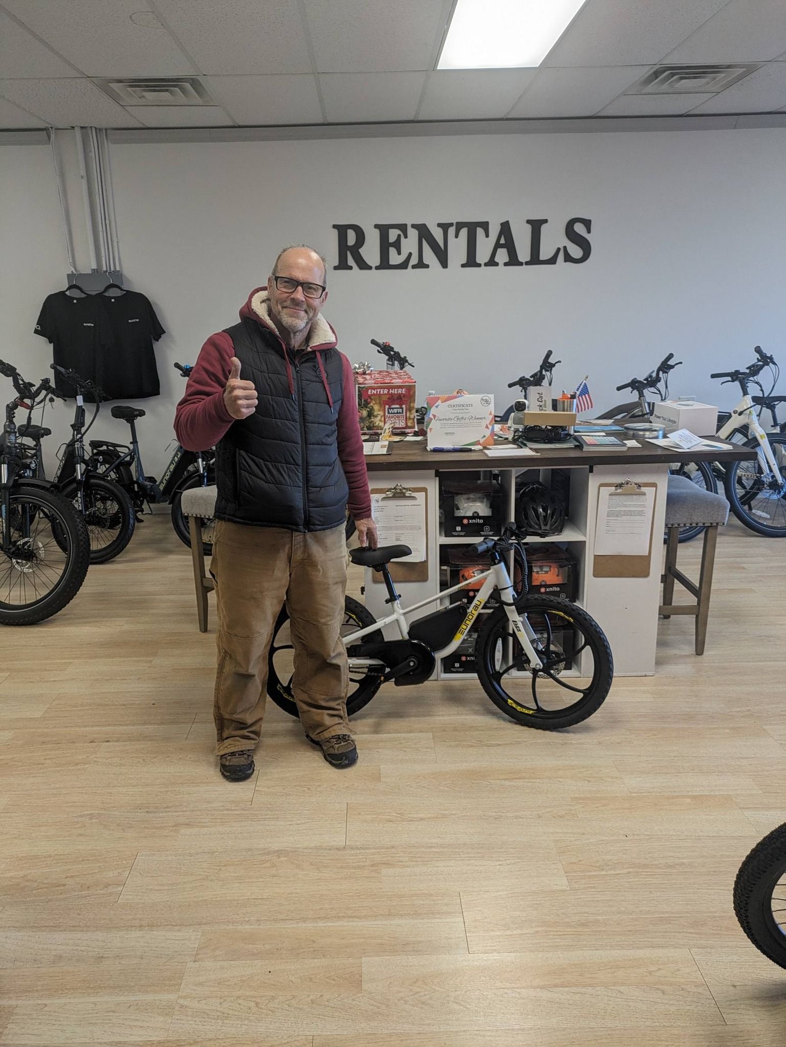 Man standing beside a black bike inside a rental shop with “RENTALS” sign on the wall