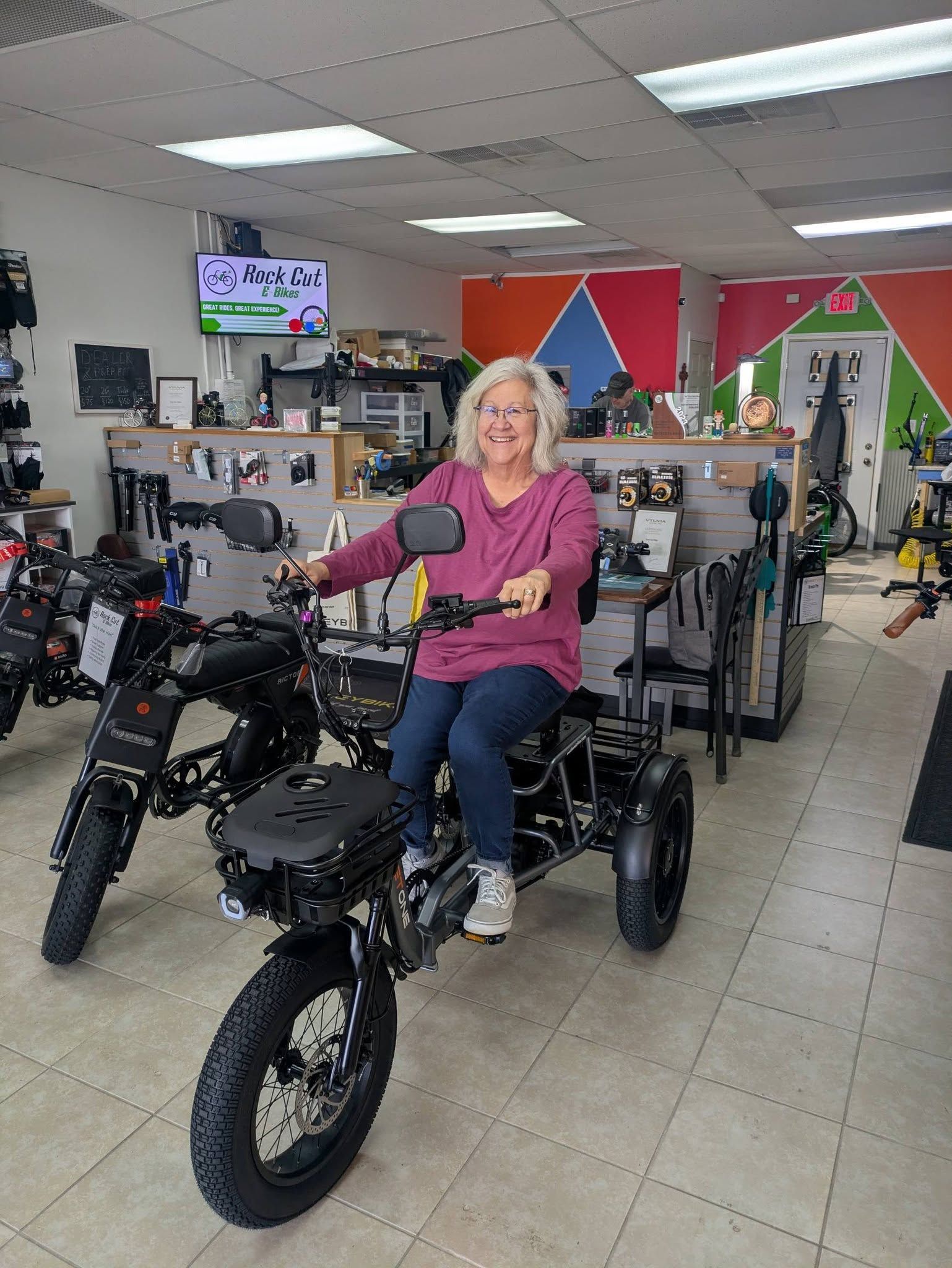 Person on a black electric trike inside a bike shop, with rows of bicycles in the background.