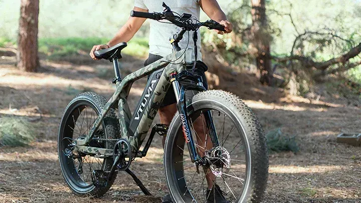 Person standing with a black mountain bike on a wooded trail