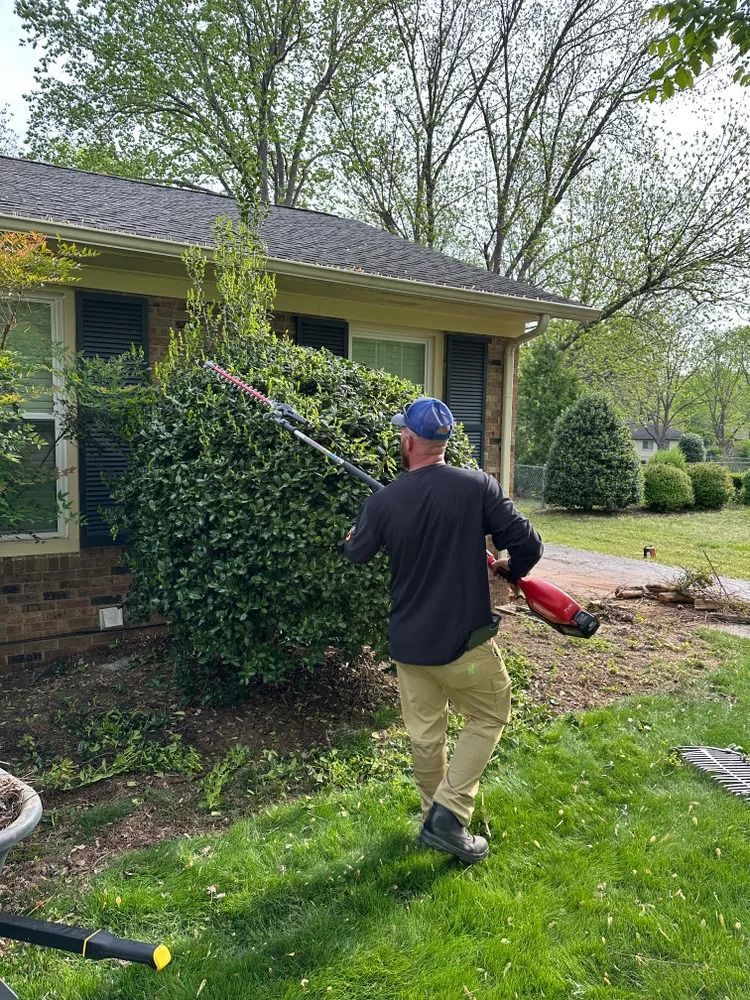 Man trimming a bush with electric shears in front of a house. Green grass, tan pants, and blue hat.