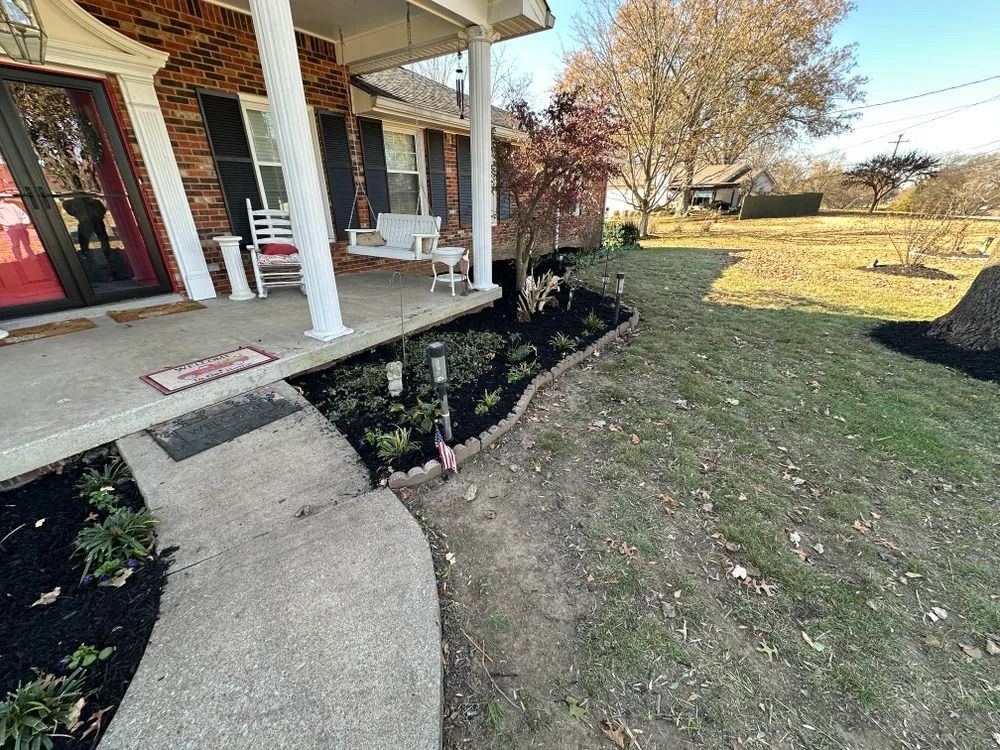 A house with a porch and a newly landscaped garden bed with black mulch and young plants.