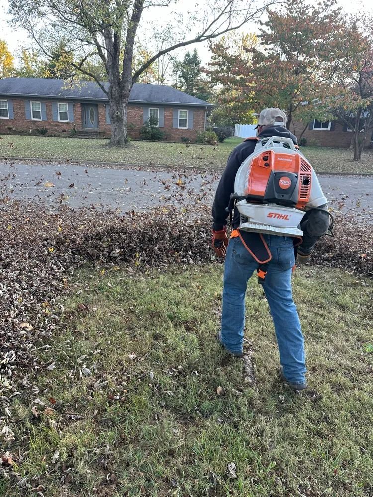 Man using a leaf blower in a yard, blowing leaves towards the street in front of a brick house.