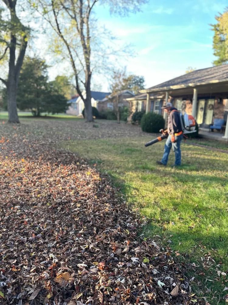 Man using a leaf blower to clear leaves from a lawn near a house on a sunny day.