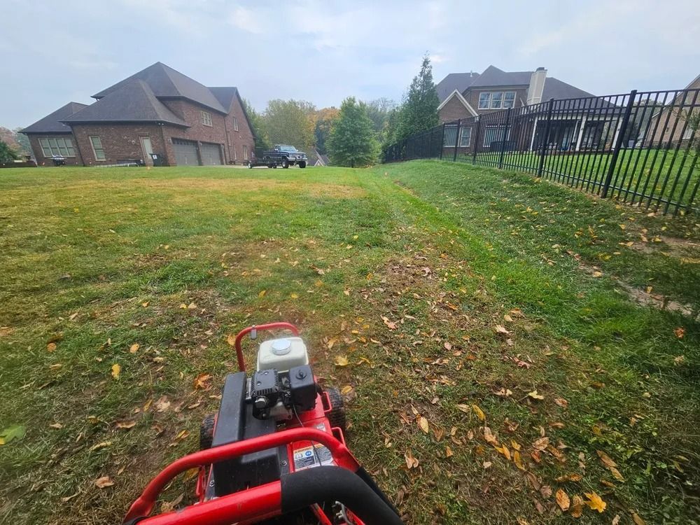 A person using a red sod cutter on a grassy hill in front of two houses on a cloudy day.