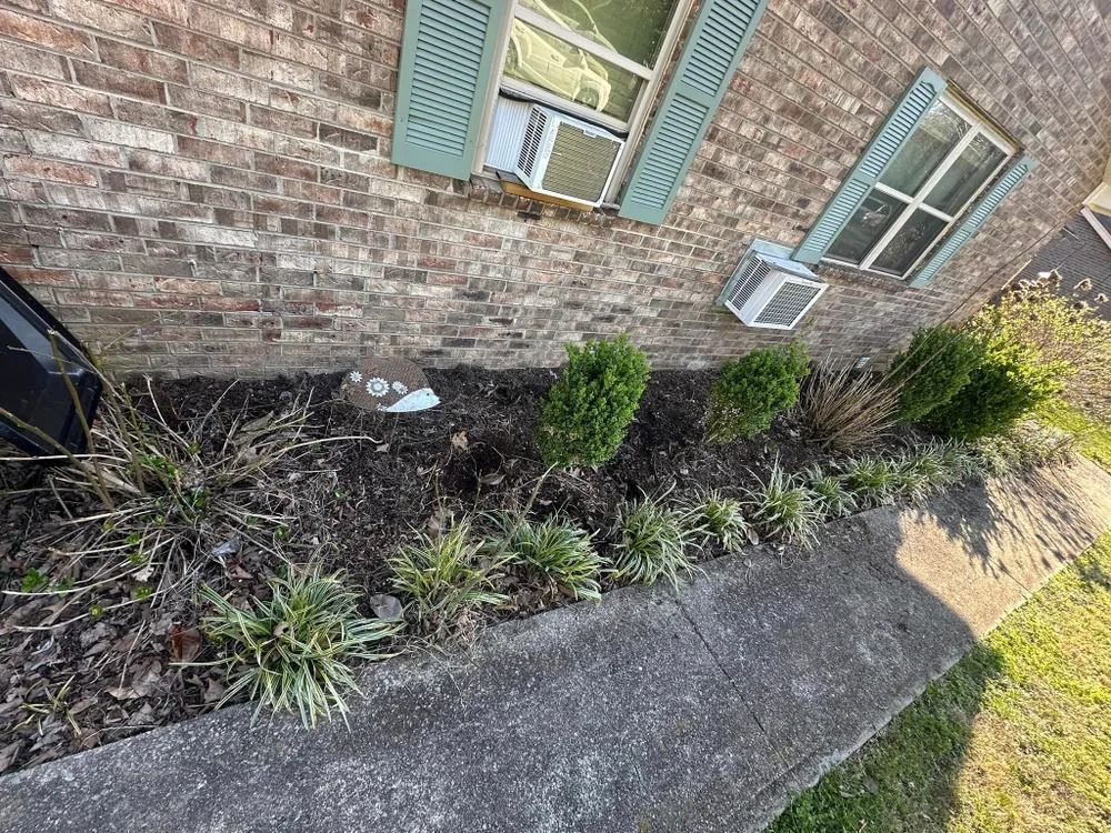Brick building with a garden bed along the sidewalk. Green shutters, bushes, and a/c units are visible.