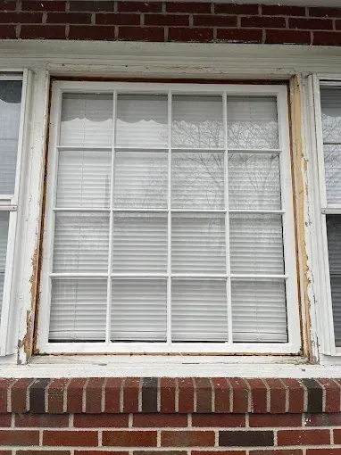 White-framed window with blinds, set in a brick wall. Wood trim shows signs of wear.