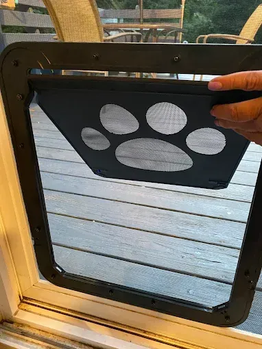 A black dog door installed in a window screen, with a paw print flap, being held open.