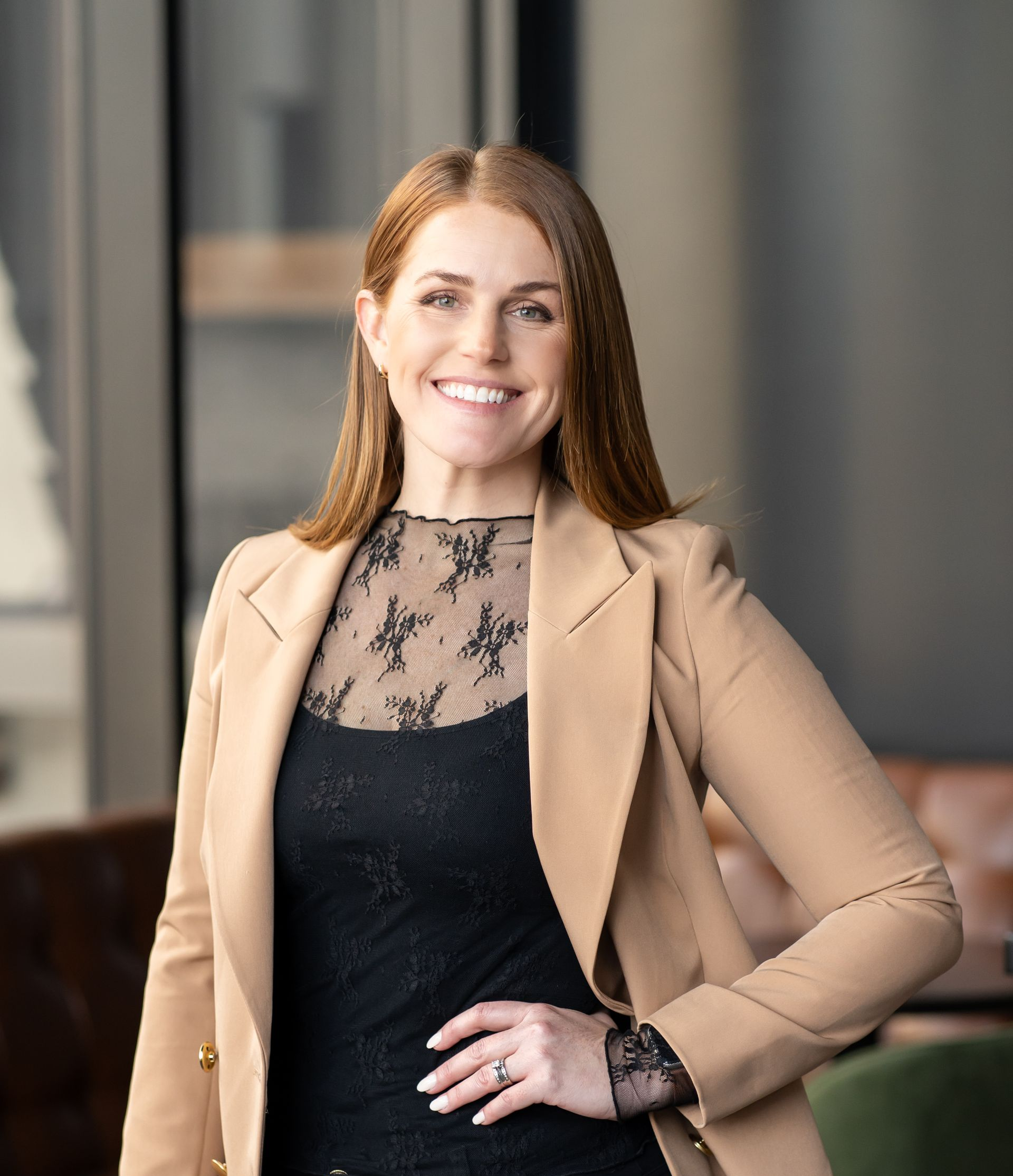 Woman in a tan blazer and black lace top smiles, arm on hip, indoors.