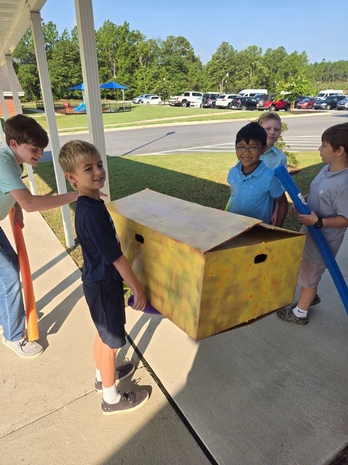 A group of young boys are carrying a large cardboard box on a sidewalk.