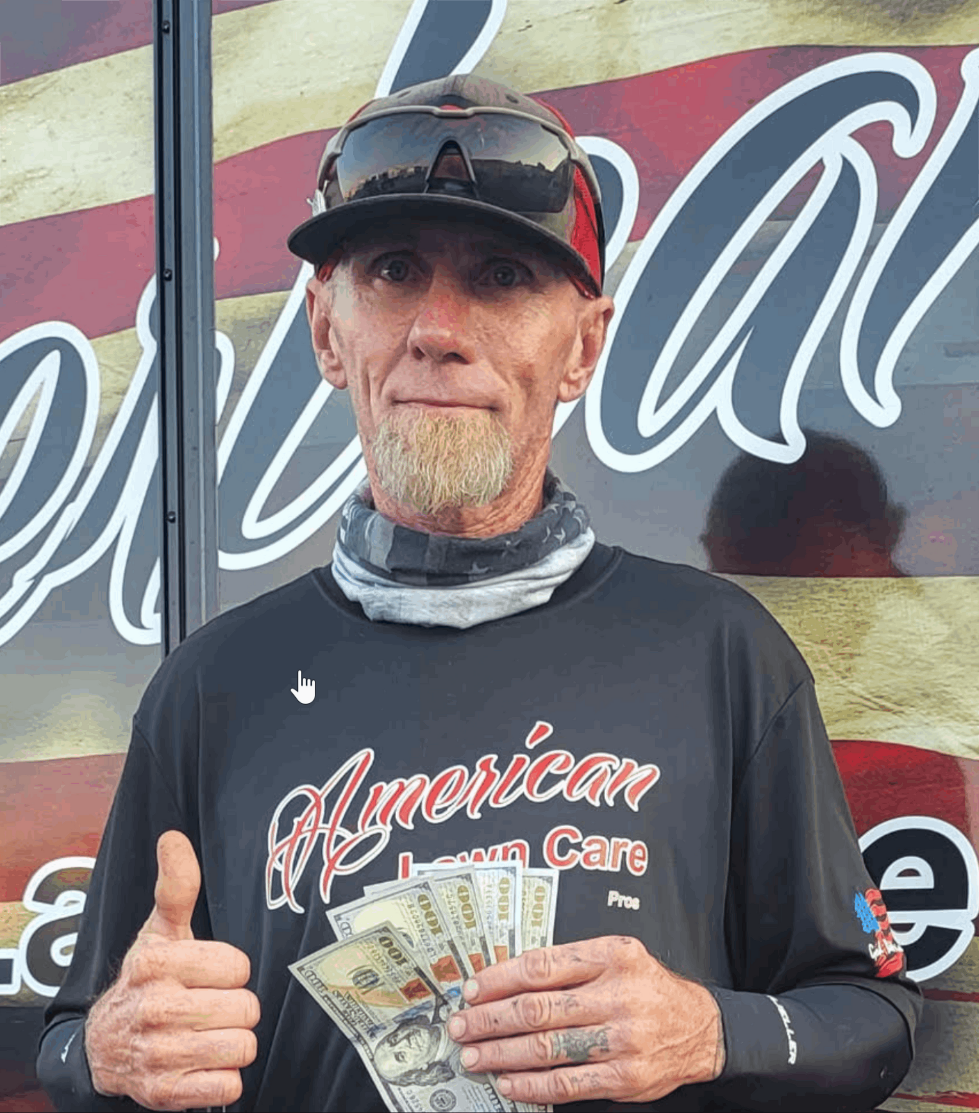A person in an American Lawn Care shirt holding a fan of hundred-dollar bills and giving a thumbs-up against a flag mural.