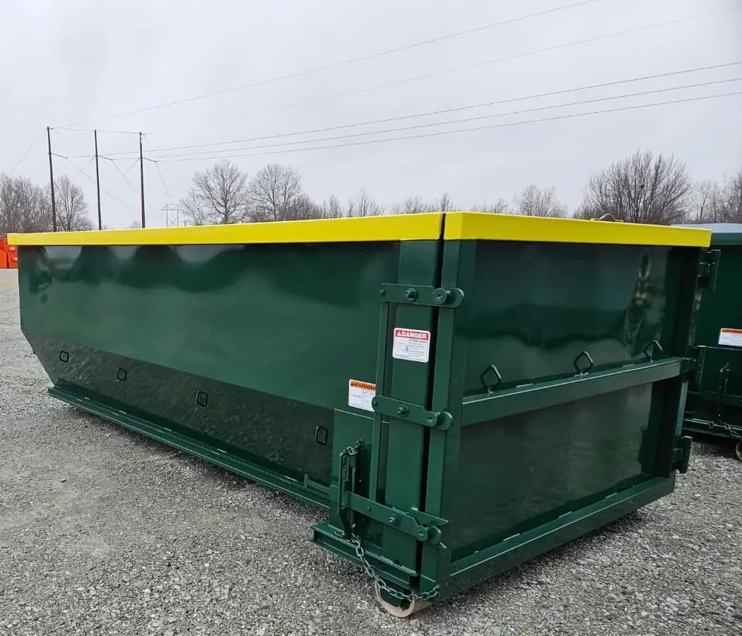 A green dumpster with a yellow lid is sitting on top of a gravel road.