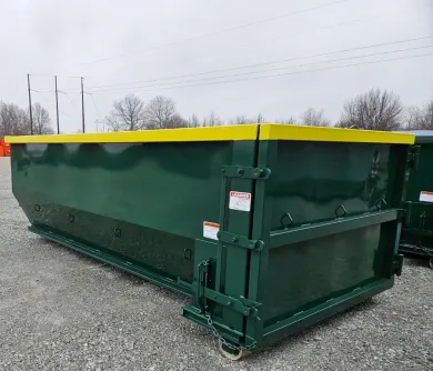 A green dumpster with a yellow lid is sitting on top of a gravel road.