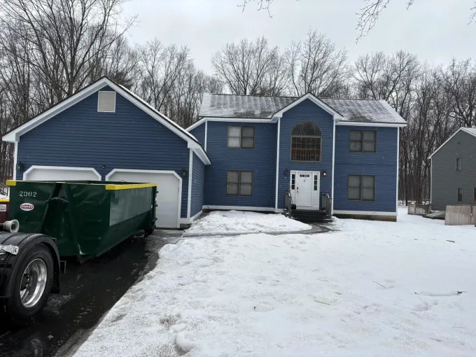 A dumpster is parked in front of a blue house covered in snow.