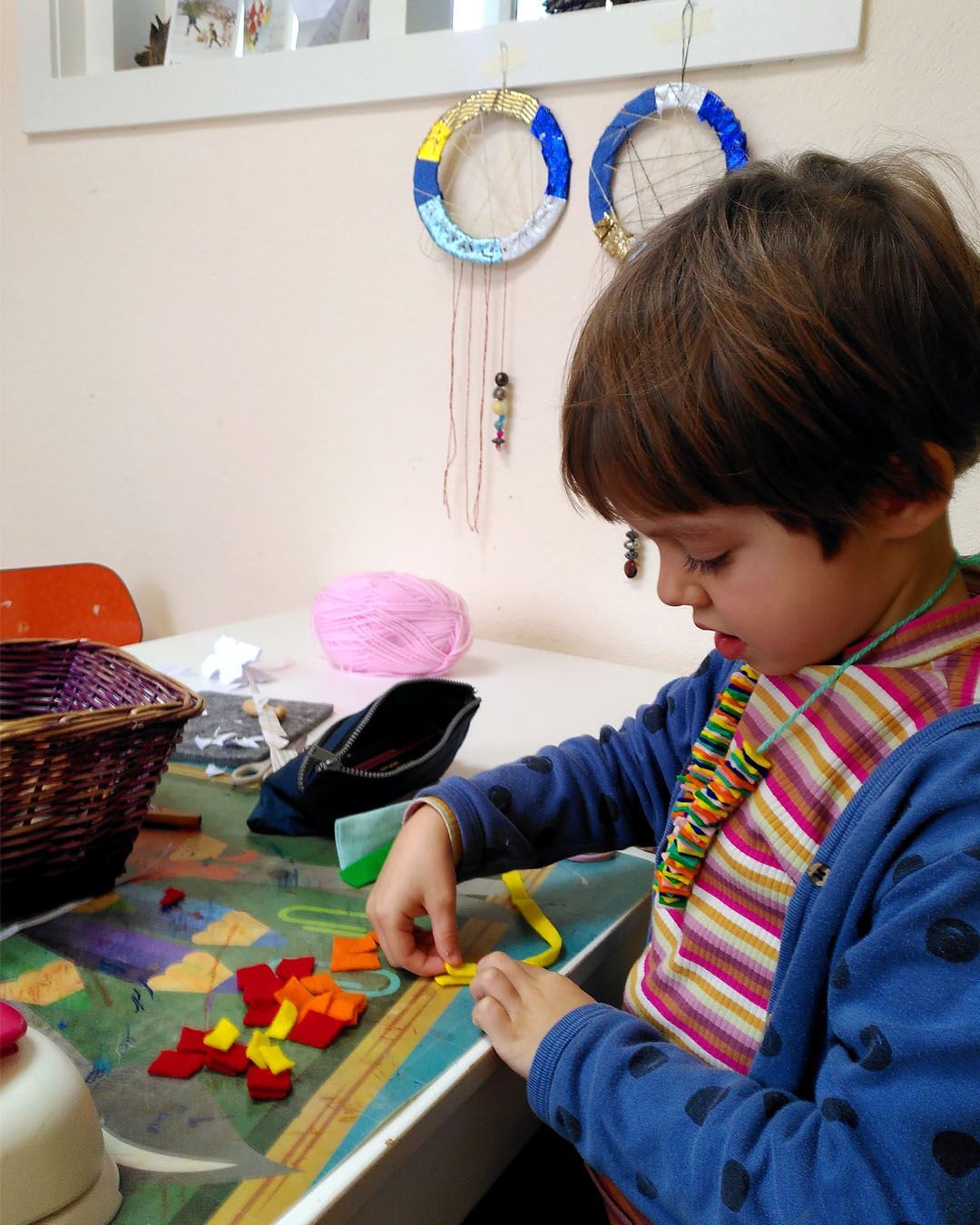 Een klein meisje zit aan een tafel en maakt met garen en vilt een ketting. Atelierkinderen, atelier Verda.