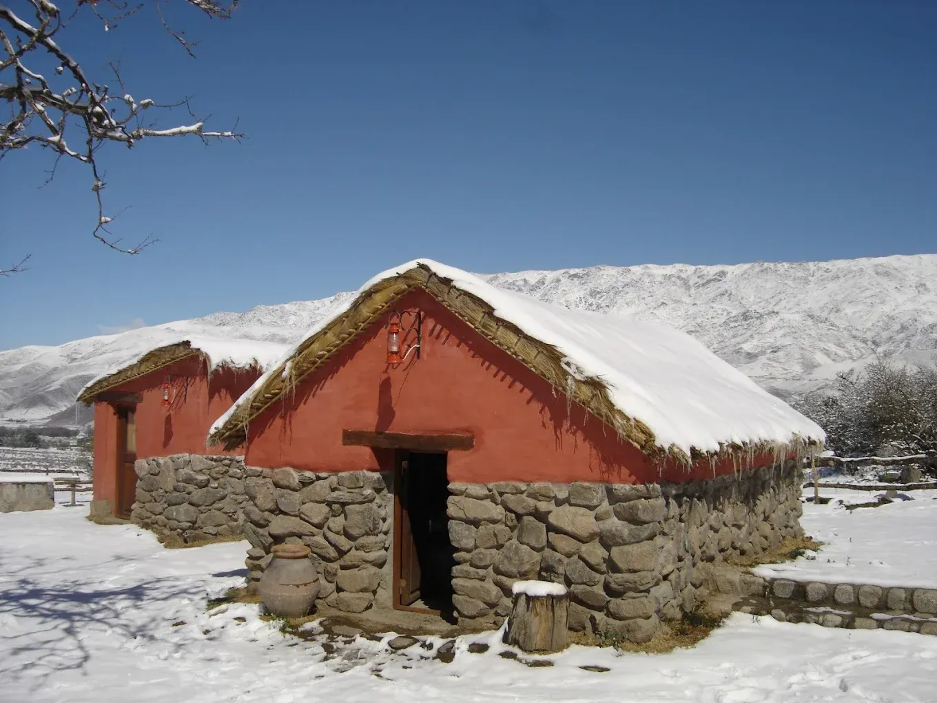 Dos cabañas de paredes rojas y techos de paja se encuentran en un paisaje nevado, con montañas al fondo bajo un cielo azul.