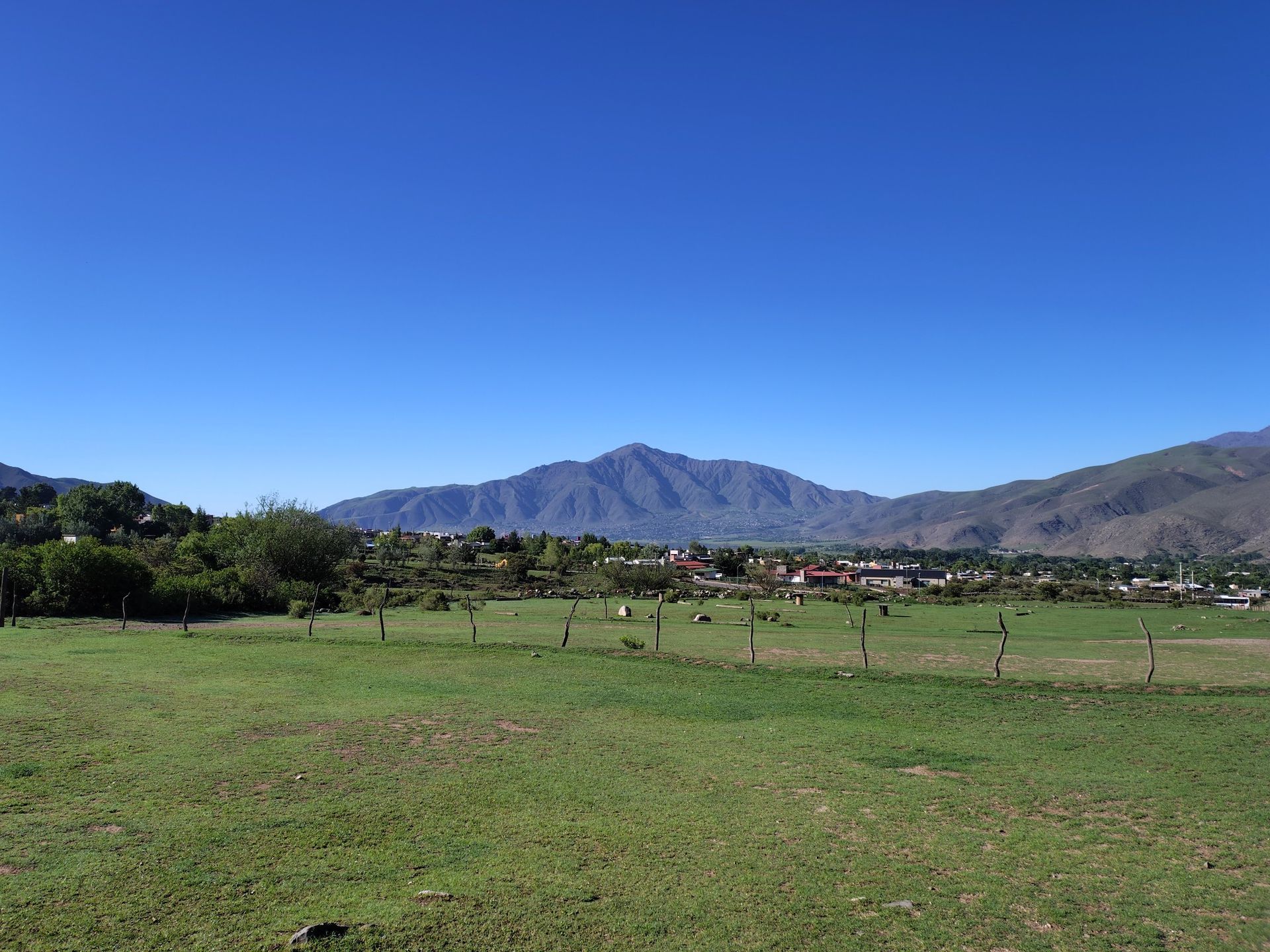 Campo verde con un pequeño pueblo al base de las montañas, bajo un cielo azul claro.