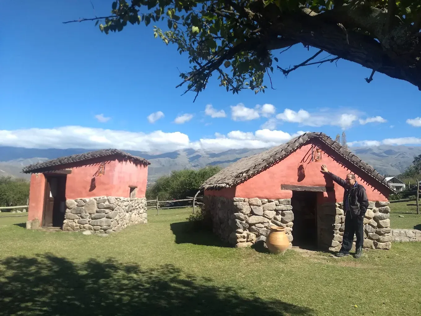 Dos casas de adobe rojo con bases de piedra y techos de paja, en un prado con montañas. 