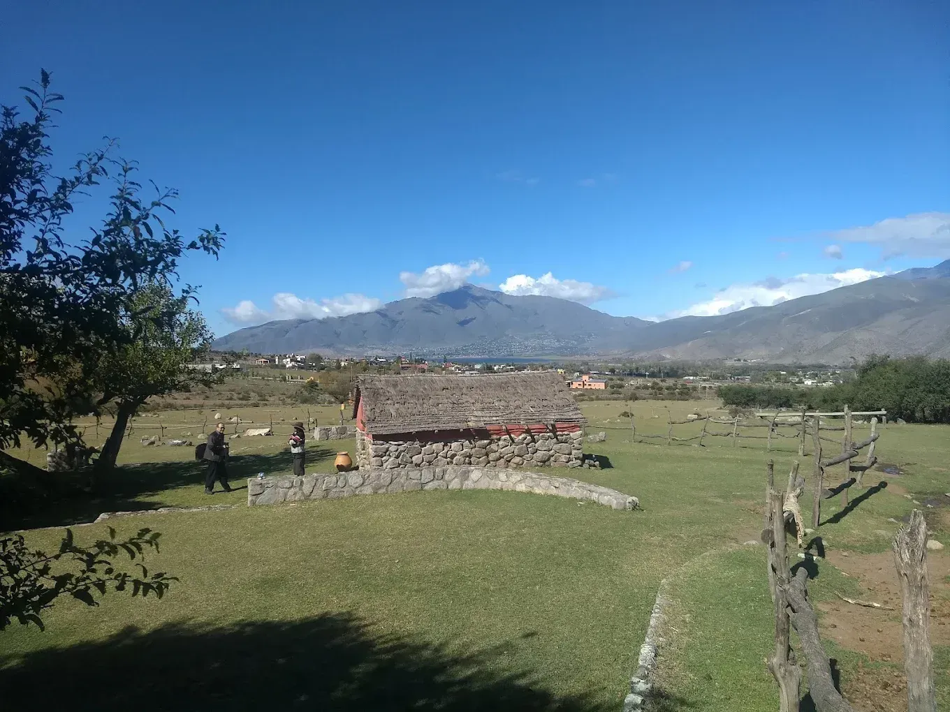 Campo de hierba con estructura de piedra, montañas y cielo azul. Hay algunas personas y una valla de madera.